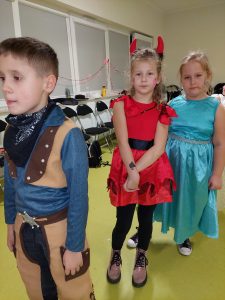 Dwie uczennice w przebraniu pozują do wspólnego zdjęcia. Przed nimi stoi chłopiec w stroju kowboja. Jedna z uczennic ma czerwoną sukienkę i czerwone rogi na głowie, a druga elegancką sukienkę w kolorze morskim.