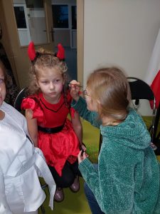 Dziewczynka w stroju diabełka siedzi na krzesełku. Przed nią kuca dziewczynka z pędzelkiem w dłoni malująca jej twarz.