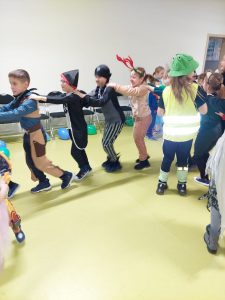 Uczniowie w soich przebraniach tańczą na zabawie andrzejkowej. Dzieci sustawione są w rzędzie i opierają ręce na ramionach kolegi stojącego przed nimi.