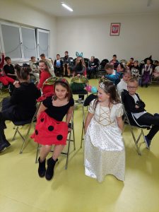 Uczniowie w swoich kostiumach siedzą na krzesełkach ustawionych po małym okręgu. W tle widać siedzące inne dzieci.