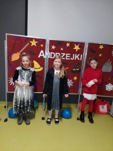 Trzy dziewczynki stoją w swoich kostiumach na tle czerwonej tablicy z napisem : "ANDZREJKI" ozdobionej kolorowymi obrazkami symbolizującymi andrzejki.