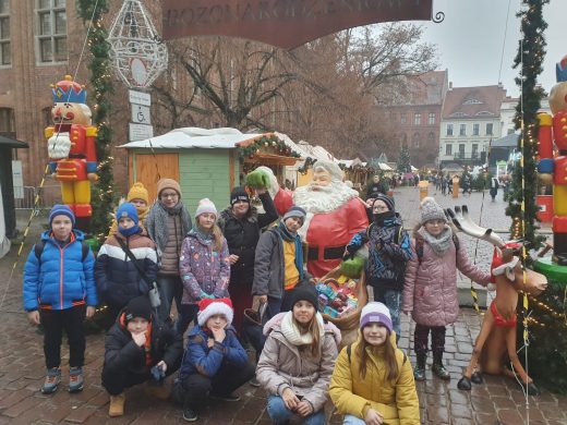 Uczniowie klasy czwartej pozują do wspólnego zdjęcia ustawieni wokół dużego Mikołaja z zieloną ręką. Część uczniów ustawionych z przodu kuca do zdjęcia. W tle widać fragment toruńskiego starego rynku.