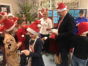 Pan wójt rozdaje uczniom czekoladki. Na glowie ma czapeczkę mikołaja. Obok stoi Pani Dyrektor a przed nimi dzieci również w czapeczkach świętego mikołaja.