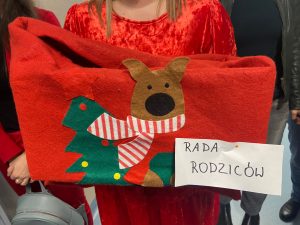 Zdjęcie trzymanej przez Panią w czerwonej sukience czerwonej torby z naklejoną choinką oraz misiem i karteczką :RADA RODZICÓW.