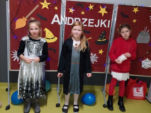 Trzy dziewczynki stoją w swoich kostiumach na tle czerwonej tablicy z napisem : "ANDZREJKI" ozdobionej kolorowymi obrazkami symbolizującymi andrzejki.