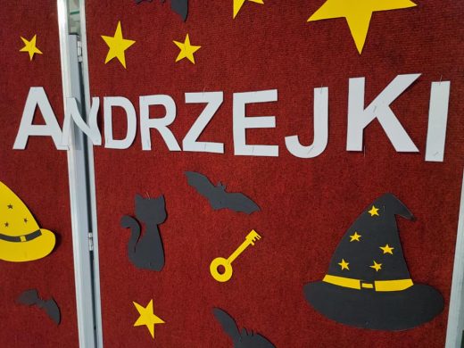 Na czerwonej tablicy biały napis "ANDZREJKI" a w okół niego wycięte z papieru dekoracje andrzejkowe takie jak: klucz, gwiazdki, kapelusz czarownika. Wszystkie ozdoby w żółtym lub czatnym kolorze.