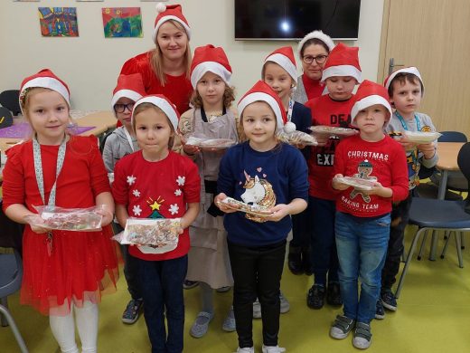 Uczniowie klasy pierwszej w czapeczkach Mikołaja pozują wraz z Panią do wspólnego zdjęcia. Dzieci trzymają w dłoniach swoje wypieki.