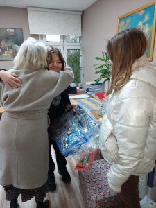 Pani w białym płaszczu obejmuje jedną z naszych uczennic w podziękowaniu za przyniesione paczki dla podopiecznych hospicjum. Obok stoi jej koleżanka a pomiędzy dziewczynkami ustawione są zapakowane prezenty.