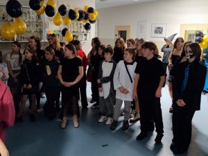 Uczniowie szkoły stoją na korytarzu. Dzieci mają na sobie różne stroje, w których przyszły na bal karnawałowy. Nad ich głowami wiszą żółte i czarne balony zawieszone w jednej linii..
