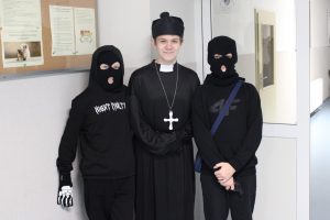 Troje uczniów klasy szóstej pozuje do wspólnego zdjęcia. Wszyscy chłopcy sa w swoich przebraniach. Po środku stoi uczeń przebrany za księdza a po jego obu stronach chłopcy w czarnych strojach i kominiarkach na twarzach.