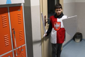 Na szkolnym korytarzu pozuje do zdjęcia uczeń klasy czwartej. Chłopiec jest w przebraniu rycerza. W jednej dłoni trzyma miecz a w drugiej podniesioną tarczę.