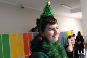 Portret ucznia stojącego na szkolnym korytarzu. Chłopiec na głowie ma opaskę ze świąteczną choinką, na uchu zawieszoną czerwoną bombkę, a na szyi zielony łańcuch choinkowy.
