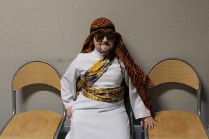 Uczeń przebrany za arabskiego szejka siedzi na krześle pomiędzy dwoma pustymi siedzeniami. Chłopiec na twarzy ma narysowany czarny zarost, na oczach duże przeciwsłoneczne okulary a na głowie czerwoną chustę spadającą na ramiona.