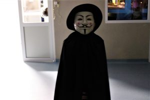 Uczeń w czarnym stroju i czarnym kapeluszu pozuje samotnie do zdjęcia na szkolnym korytarzu. Chłopiec na twarzy ma białą maskę z narysowanymi czarnymi wąsami i wąską brodą.