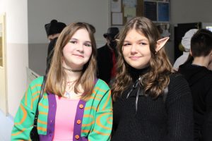 Dwie uczennice klasy ósmej pozują do wspólnego zdjęcia. Uczennica po prawej stronie ubrana na czarno ma doczepione elfie uszy. Obok niej stoi dziewczynka w kolorowym ubraniu.