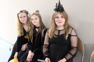 Trzy uczennice klasy czwartej pozują do wspólnego zdjęcia siedząc na krzesełkach ustawionych pod ścianą. Uczennica siedzą z przodu ma na głowie opaskę z czarnym kapeluszem wiedźmy a na twarzy namalowanego pająka. Ubrana jest w czarna sukienkę. dziewczynki siedzące za nią również ubrane na czarno mają na głowach opaski.