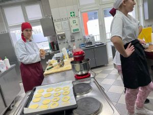 Zdjęcie wykonane w kuchni. Na pierwszym planie widać przygotowane do pieczenia ciastka rozłożone na blaszcze przykrytej białym papierem. Dalej stoi robot kuchenny a za długim blatem chłopak w stroju kucharza w czerwonej chuście na głowie. Przed stołem widać jeszcze jedną postać stojącą bokiem z ramionami opartymi na biodrach.