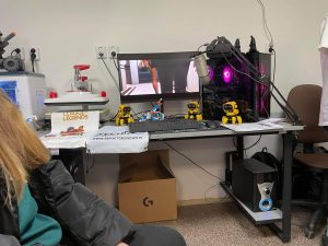 Zdjęcia biurka w jednej z sal Szkoły w Gronowie. Na biurku ustawiony jest komputer z monitorem i klawiaturą. Wokół nich poustawiano żółte roboty. Na monitorze wyświetlona jest scena z gry komputerowej.