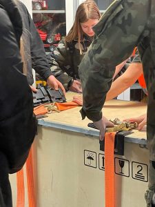 Na zdjęciu widoczne są fragmenty kilku postaci a wśród nich uczennicy klasy ósmej. Wszystkie osoby stoją wokół dużej drewnianej skrzyni i przypinają na niej pomarańczowe pasy.