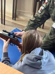 Uczennice w pozycji leżącej trzyma w dłoni broń strzelecką. Obok stoi instruktor również podtrzymujący broń. Na zdjęciu widać tył głowy uczennicy i fragment jej bluzy.
