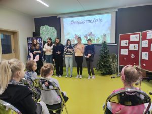 Sześcioro uczniów klasy szóstej stoi w rzędzie na sali teatralnej Dwoje z nich trzyma w dłoni mikrofony. Po prawej stronie widać fragment czerwonej tablicy z przyczepionymi kartkami i śnieżynkami, a za uczniami na białym ekranie wyświetla się grafika z napisem Bezpieczne ferie i w górę skierowanymi kolorowymi dłoniami. Przed uczniami na krzesełkach siedzą dzieci oglądające prezentację.