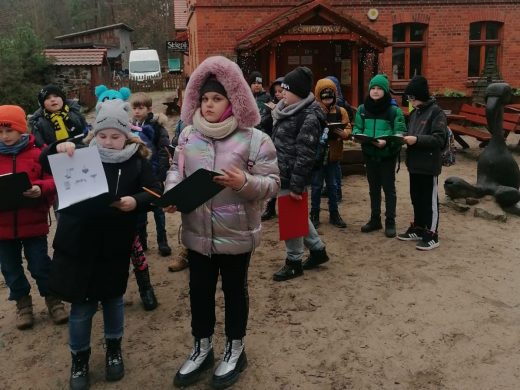 Uczniowie stoją w kurtkach przed budynkiem leśnej szkoły na Barbarce. Dzieci trzymają w dłoniach podkładki z kartkami.