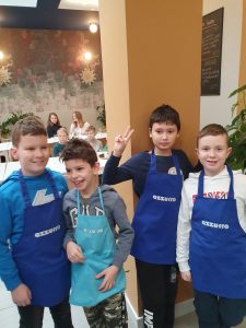 Czterech uczniów klasy czwartej pozuje do wspólnego zdjęcia w niebieskich fartuchach kuchennych z napisem azzurro. Jeden chłopiec podnosi w górę rękę z wyprostowanymi dwoma palcami. W tle widać siedzących przy długich stołach innych uczniów.