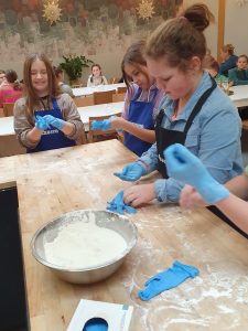 Uczennice klasy czwartej stoją przy wysokim stole z drewnianym blatem. Stół posypany jest mąką. Stoi na nim metalowa miska wypełniona mąka oraz pudełko z lateksowymi rękawiczkami. Dziewczynki zakładają rękawiczki na swoje dłonie.