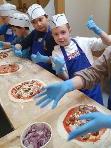 Czworo chłopców klasy czwartej stoi przy wysokim stole z drewnianym blatem. Chłopcy na głowie mają papierowy czapeczki a na dłoniach niebieskie, lateksowe rękawiczki. Dwóch z nich podnosi w górę kciuki. Przed chłopcami na stole leżą przygotowane przez nich pizze.