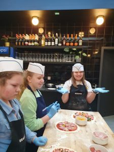Trzy uczennice klasy czwartej stoją przy wysokim, drewnianym blacie. Dziewczynki na głowach mają białe, papierowe czapeczki, a na dłoniach niebieskie, lateksowe rękawiczki. Uczennice stojąca na szczycie stołu szeroko się uśmiecha i rozkłada szeroko dłonie. Na stole przed dziewczynkami leżą przygotowywane przez nie pizze oraz białe miseczki ze składnikami.