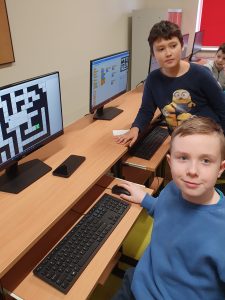 Uczeń siedzi przy komputerze. Odwraca głowę w stronę zdjęcia. Przed nim na monitorze wyświetla się ekran z czarno-białą planszą gry. Za nim stoi jego kolega. Chłopiec patrzy w bok.