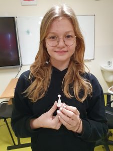 Uczennica uśmiecha się. W dłoniach przed sobą trzyma wydrukowany w drukarce 3D pionek do gry.