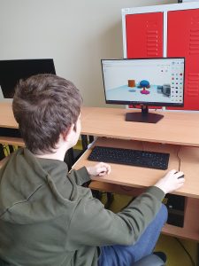 Uczeń siedzi przy komputerze. Na ekranie przed nim widać projekt pionka i kostki do gry, nad którymi pracuje uczeń.