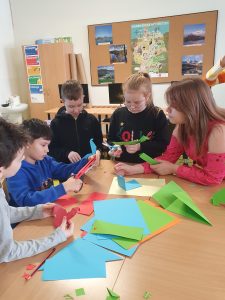 Uczniowie podczas wspólnej pracy nad origami. Dzieci siedzą lub stoją przy stoliku, na którym ułożono kolorowe kartki.