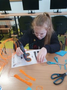 Uczennica siedzi przy stole i wykonuje pomarańczowe okulary przy użyciu długopisu 3D. Przed uczennica na stole lerzą gotowe juz elementy oraz nozyczki. Dziewczynka przyciska długopis 3D do kartki, na której ma odrysowany schemat okularów.