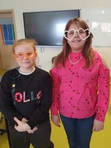 Dwie uczennice pozują do wspólnego zdjęcia. Dziewczynki mają założone na twarzy wykonane przez siebie okulary. Dziewczynka stojąca po lewej stronie wykonała pomarańczowe okulary w klasycznym kształcie, a ta stojąca po prawej wykonała białe okulary w kształcie serc.