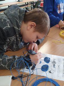 Uczeń podczas pracy z długopisem 3D. Chłopiec pochyla się nad kartką ze schematem elementów potrzebnych do złożenia okularów. W dłoni trzyma długopis 3D z niebieskim wkładem.