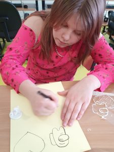 Uczennica pochyla się nad kremową kartką na której rysuje wzory do wykonania długopisem 3D. Obok niej stoi wydrukowany w drukarce 3D pionek w kształcie dwóch misiów stojących na okrągłym podeście.