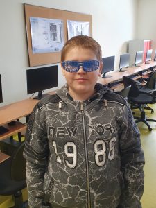 Uczeń pozuje do zdjęcia. Na twarzy ma założone wykonane przez siebie niebieskie okulary. Okulary zamiast szkieł mają niebieską kratkę.