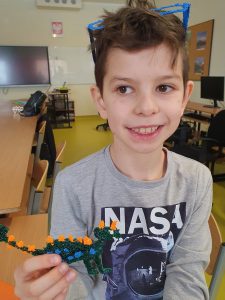 Uczeń uśmiecha się. Na głowie ma założone niebieskie okulary, które wykonał przy użyciu długopisu 3D, a w prawej dłoni trzyma wykonanego przez siebie ta są techniką dinozaura.
