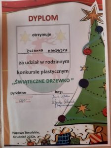 Zdjęcie dyplomu otrzymanego przez Zuzannę Makowską za udział w rodzinnym konkursie plastycznym "ŚWIĄTECZNE DRZEWKO". Dyplom ozdobiony grafiką udekorowanej choinki i ułożonymi pod nią prezentami.