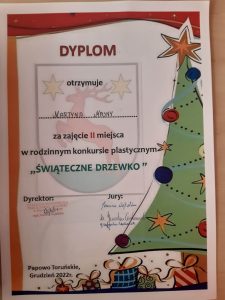 Zdjęcie dyplomu otrzymanego przez Martynę Masny za zajęcie II miejsca w rodzinnym konkursie plastycznym "ŚWIĄTECZNE DRZEWKO". Dyplom ozdobiony grafiką udekorowanej choinki i ułożonymi pod nią prezentami.