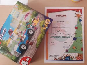 Zdjęcie dyplomu i nagrody otrzymanej przez Martynę Masny za zajęcie drugiego miejsca w rodzinnym konkursie plastycznym "ŚWIĄTECZNE DRZEWKO". Po prawej stronie ułożono dyplom a po lewej częściowo na niego nachodzące pudełko z klockami lego.