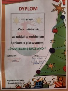 Zdjęcie dyplomu otrzymanego przez Lenę Sadowską za udział w rodzinnym konkursie plastycznym "ŚWIĄTECZNE DRZEWKO". Dyplom ozdobiony grafiką udekorowanej choinki i ułożonymi pod nią prezentami.