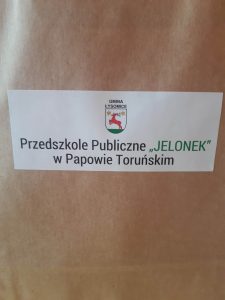 Na brązowym tle biała naklejka z logiem gminy Łysomice i napisem "Przedszkole Publiczne "Jelonek" w Papowie Toruńskim.