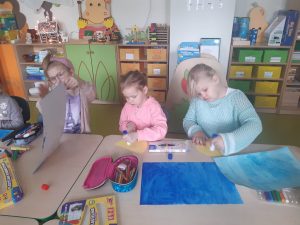 Trzy uczennice siedzą przy stole i wykonują swoje prace plastyczne przedstawiające układ słoneczny. Na stoliku przed dwiema uczennicami smarującymi klejem żółte półkola, leżą pomalowane na niebiesko kartki papieru oraz przybory plastyczne. Dziewczynka siedząca obok nich podnosi w górę swoją pracę.