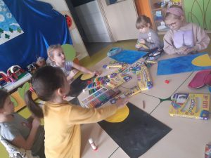Uczniowie oddziału przedszkolnego podczas wykonywania prac plastycznych przedstawiających układ słoneczny. Przy dużym stole siedzą dziewczynki. Przed każdą z nich leży częściowo wykonana praca. Pomiędzy pracami leżą rozłożone przybory plastyczne.
