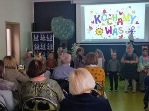 Uczniowie oddziału przedszkolnego podczas wystąpienia z okazji dnia babci i dziadka. Na pierwszym planie widać siedzących gości a przed nimi w rządku stoją dzieci w swoich strojach z przedstawienia. Za dziećmi na białym tle wyświetlony jest kolorowy napis "KOCHAMY WAS" udekorowany grafikami kwiatków i motylków.