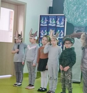 Pięcioro uczniów oddziału przedszkolnego podczas wystąpienia z okazji dnia babci i dziadka. Dzieci stoją w jednym rzędzie. Stojące od lewej strony dziewczynki na głowie mają opaski z przyczepionymi długimi uszami. Pierwsza dziewczynka trzyma mikrofon. Obok dziewczynek stoi chłopiec w żołnierskim przebraniu.