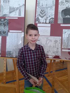 Wyróżniony w konkursie uczeń - Karol Cebulski stoi na tle wyróżnionych prac plastycznych. Chłopiec uśmiecha się.